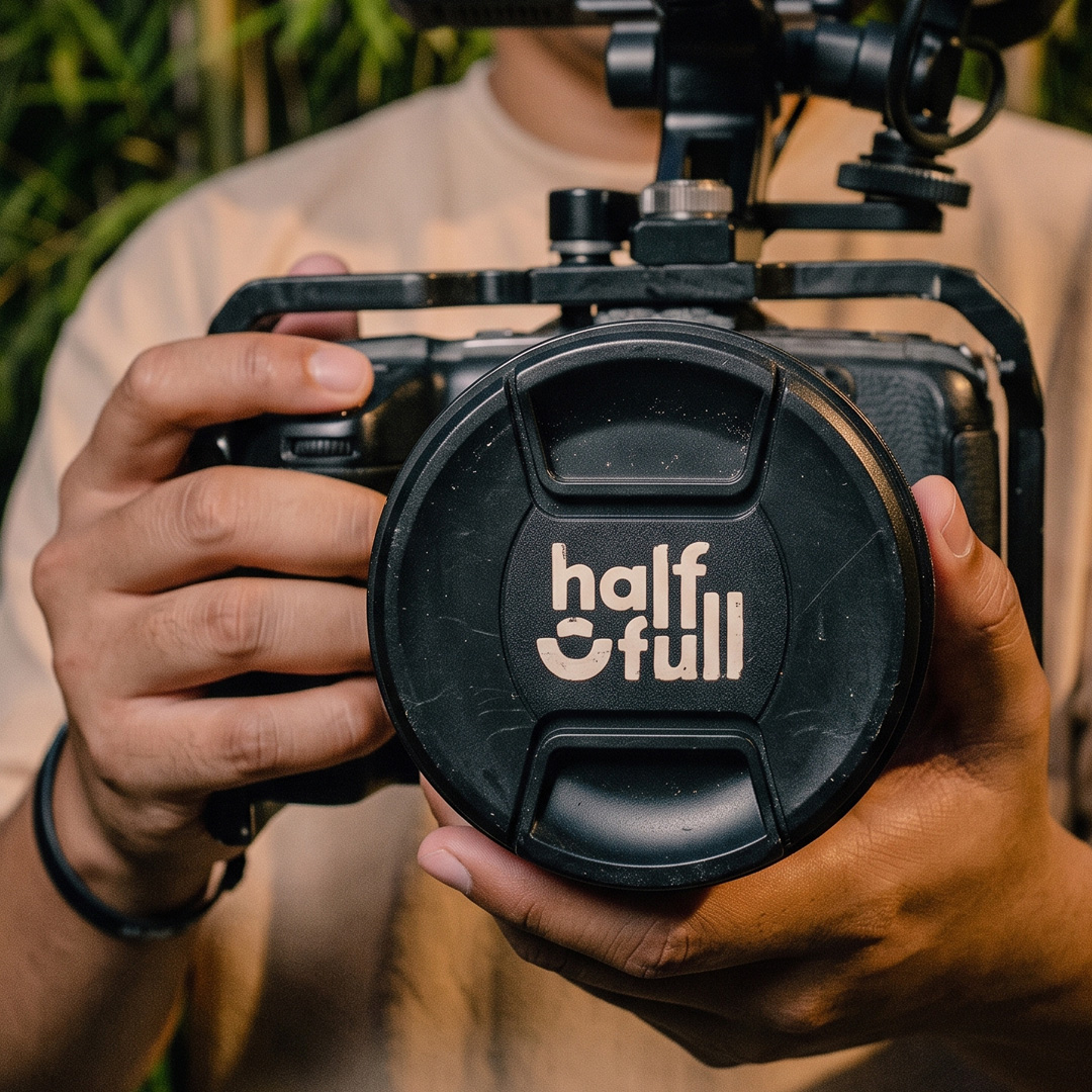 Close-up photo of hands holding a camera rig at chest level, focused on a black lens cap featuring the white "half full" logo under warm, direct flash lighting against blurred bamboo foliage.