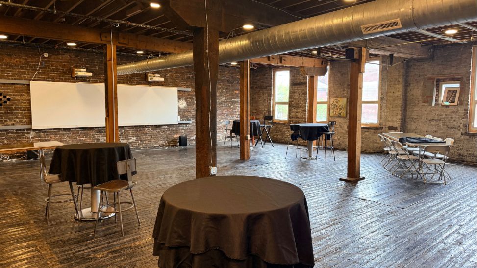 Rustic event space with exposed brick walls, wooden floor and beams, industrial ceiling ductwork, small round tables covered in black cloth, and folding chairs arranged around some tables near windows.