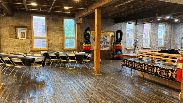 Interior of a rustic event space with wooden floors, exposed brick walls, and wooden beams on the ceiling, set up with folding chairs around black-covered tables and decorations including balloon columns shaped as the numbers 6 and 0 beside a door.