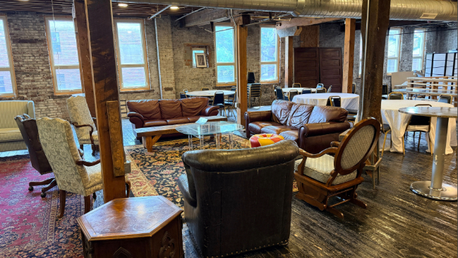 Spacious event space with exposed brick walls and wooden beams, furnished with brown leather sofas, assorted chairs, a glass coffee table on patterned rugs, and several white-draped round tables near large windows.