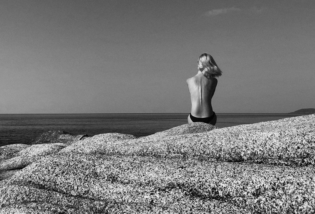 Donna di spalle seduta su rocce, che guarda l'orizzonte marino in bianco e nero