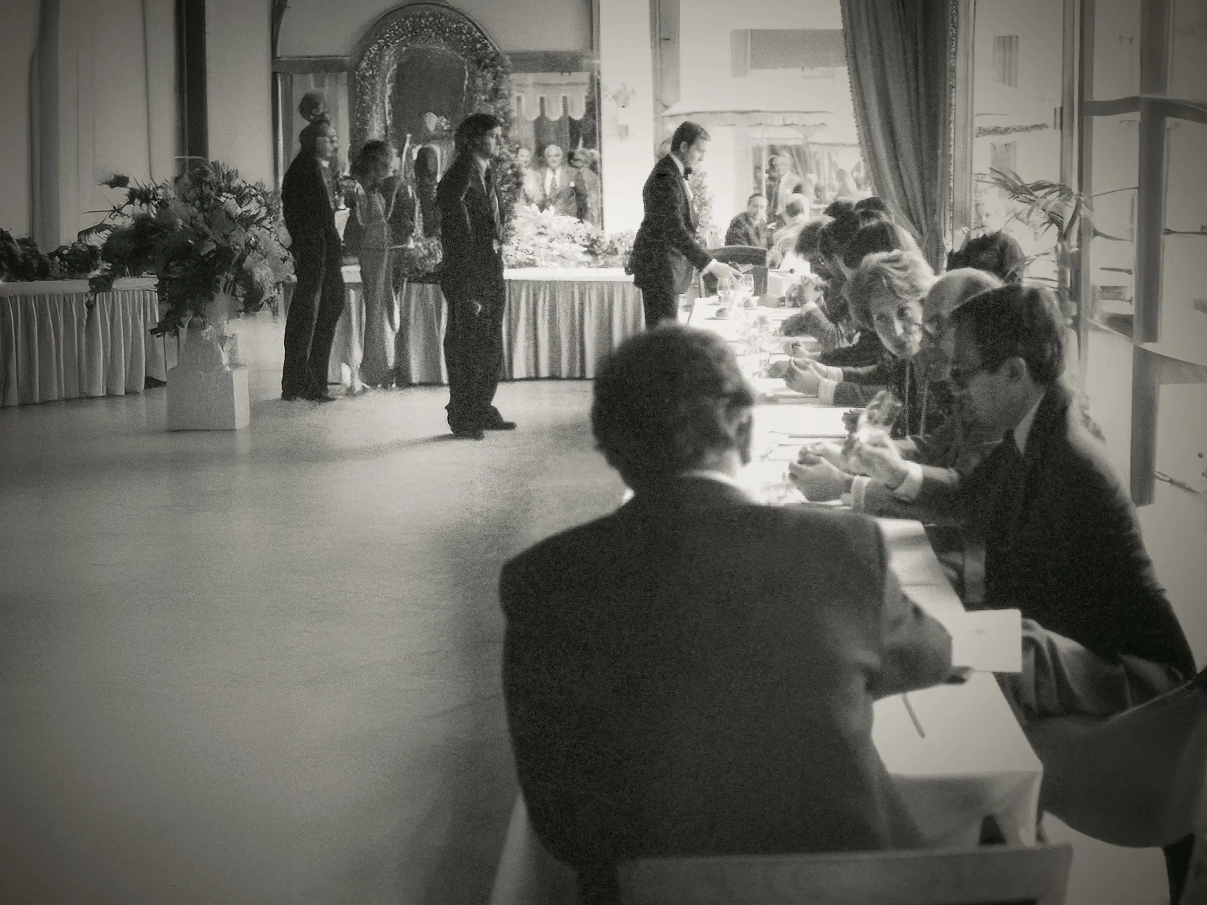 A black-and-white image of a formal event with people seated at a long table in a large, elegant room.