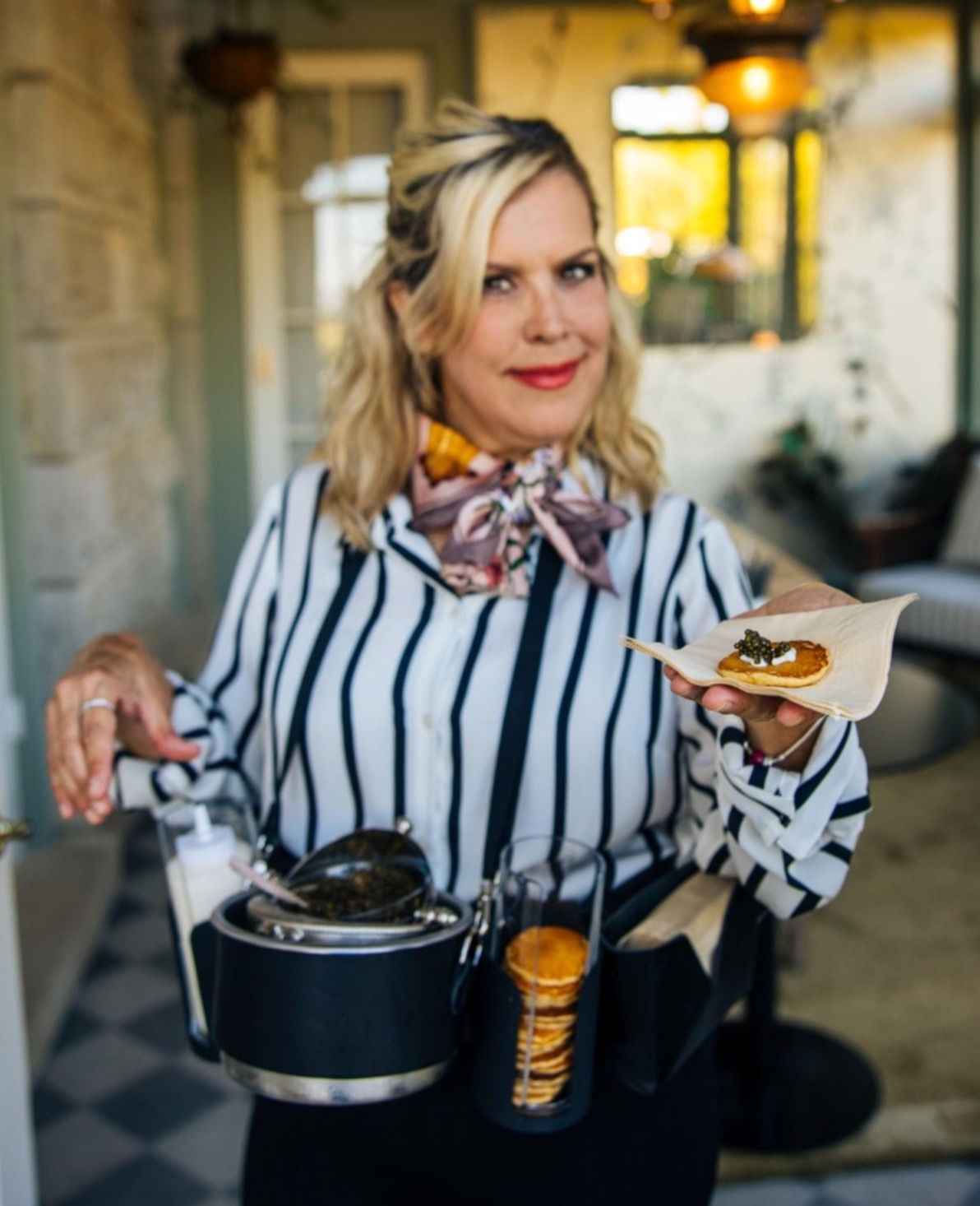 Smiling server in a striped blouse and neck scarf presenting a blini topped with caviar. A serving harness caviar, blinis and other accompaniments for guests.