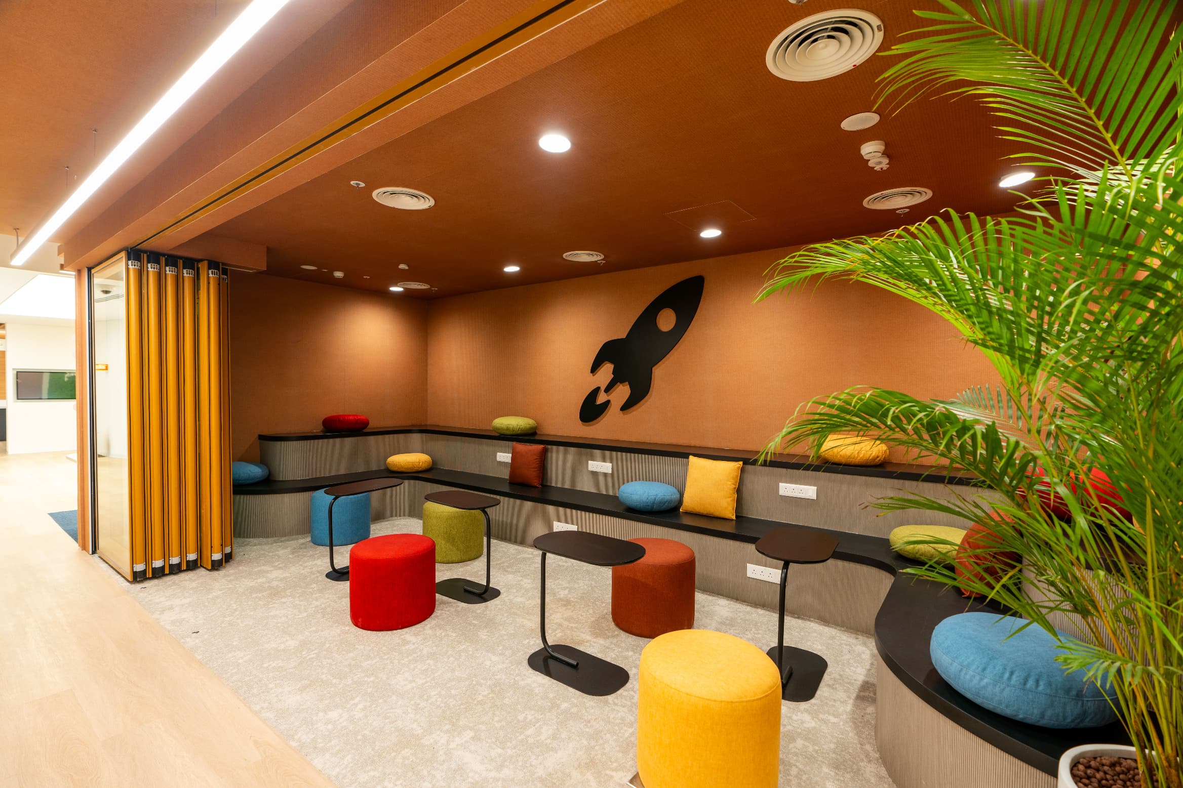 A compact pantry doubles as amphitheatre seating with agile furniture. The rocket mascot recurs here as a wall graphic, one of several points where the brand enters the room as form rather than signage.