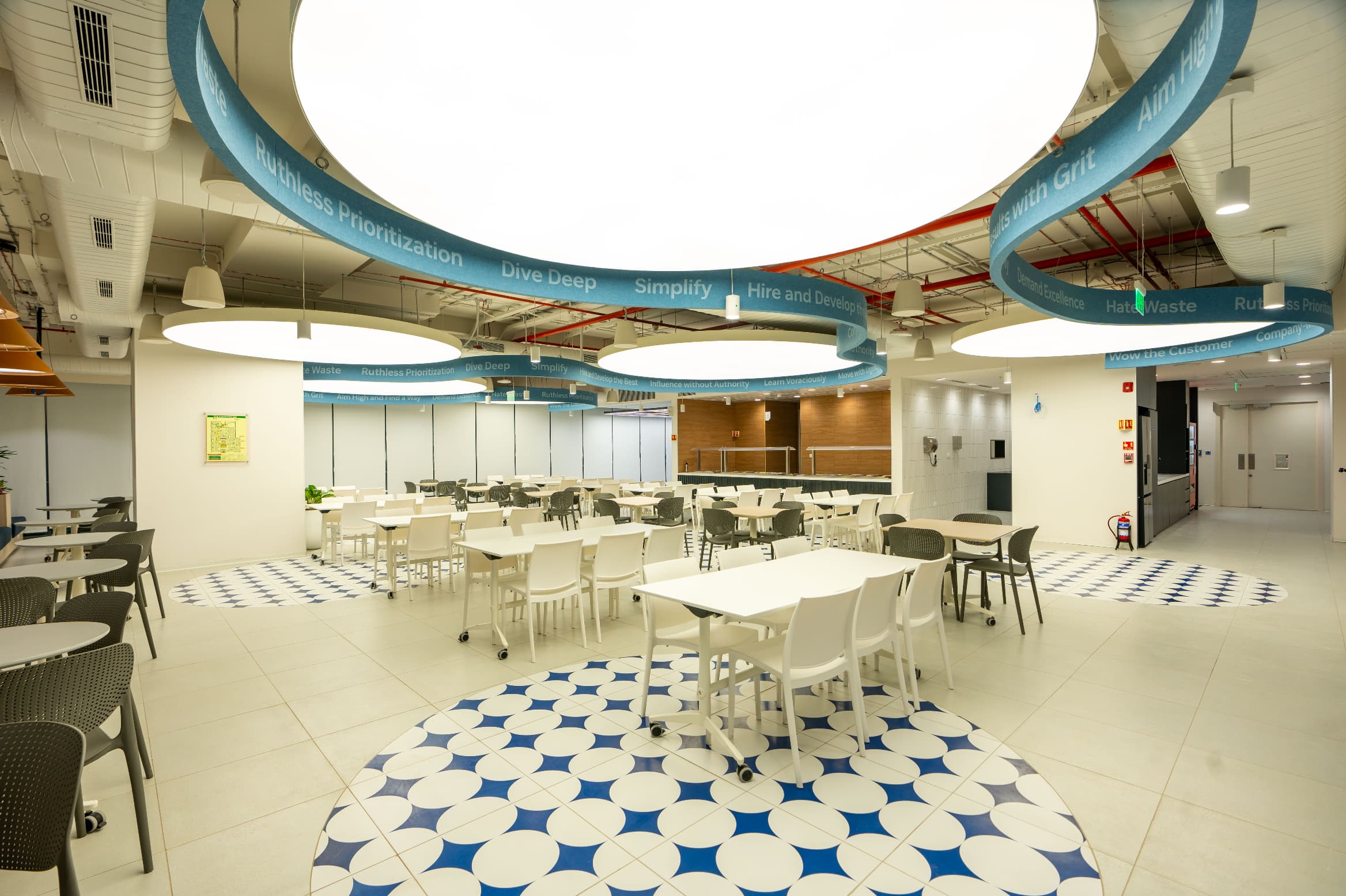 A sculptural ceiling of stretched fabric ribbons across the café carrying the company's values and vision statements. The same ribbon language is etched into the floor tiles, and at night the illuminated ceiling reads from the building façade.