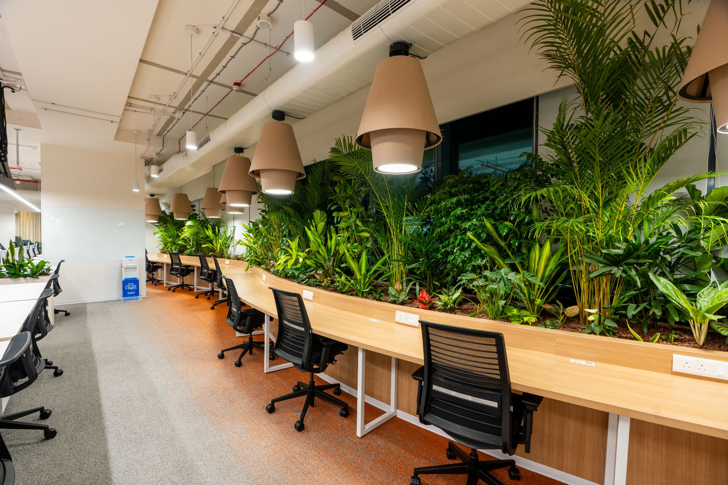 Landscaped edges run the length of the workstation banks, with planting beds built into the desk line and sculptural pendants above.