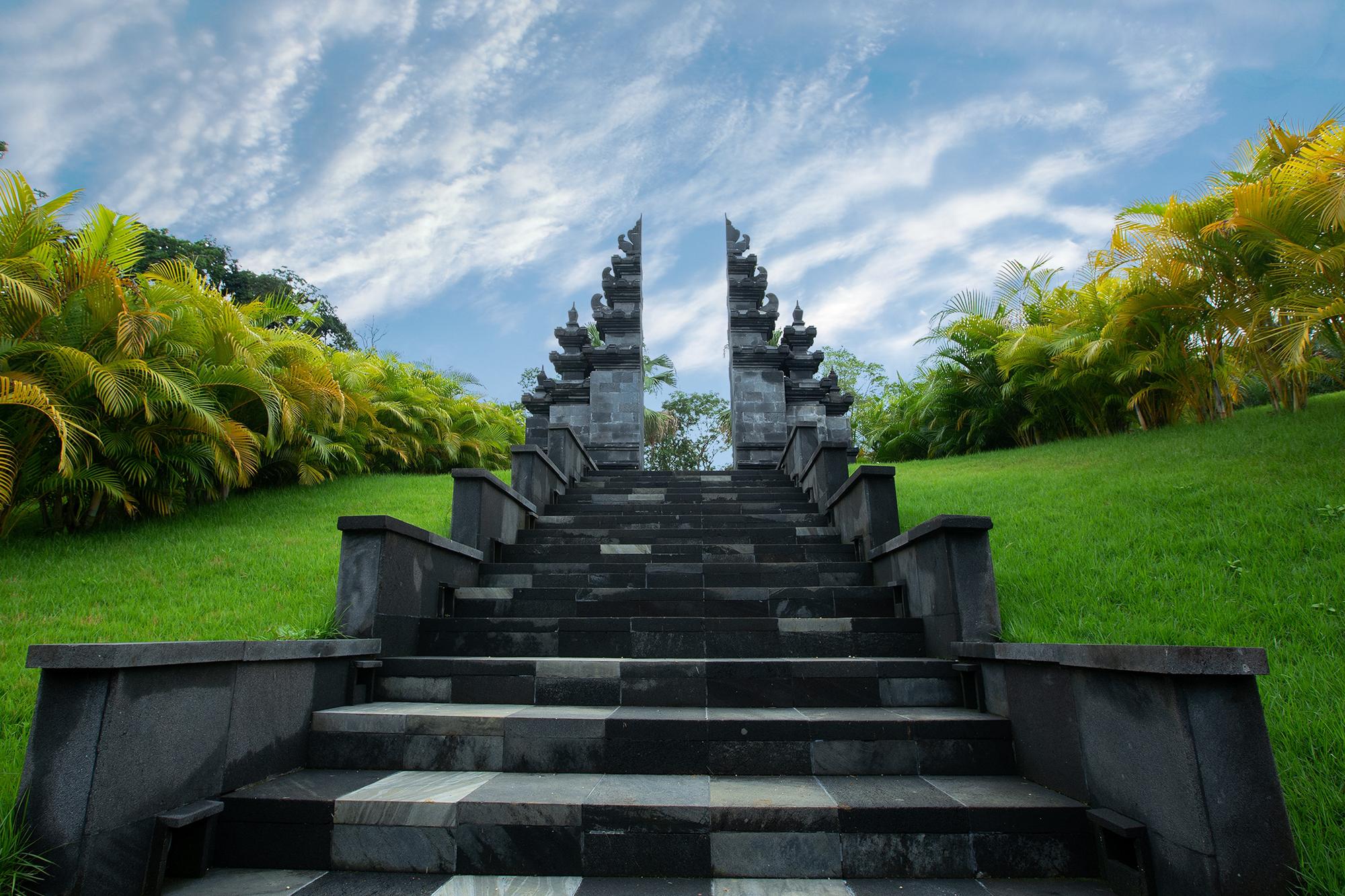 Stone staircase leading up to a traditional Balinese split gate, flanked by green grass and tropical plants under a blue sky with scattered clouds.