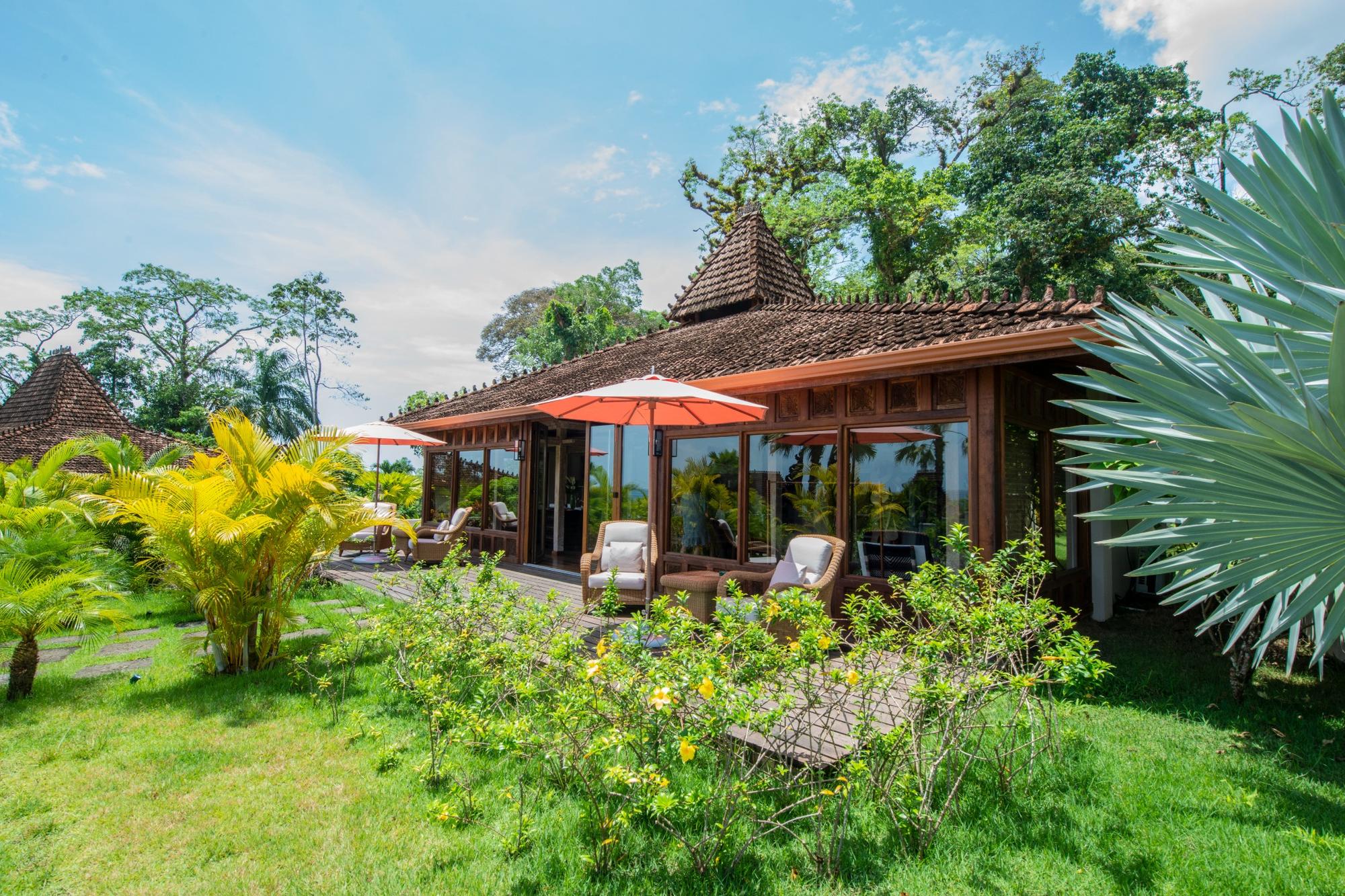 Traditional wooden house with tiled roof and large glass windows surrounded by lush green garden and patio with seating and umbrellas.