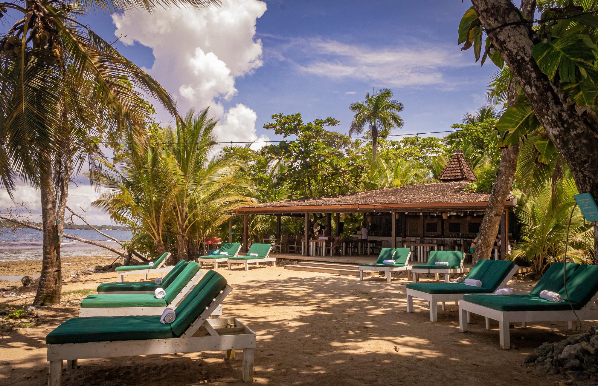 Beachfront lounge area with green cushioned sunbeds and a shaded bar hut surrounded by palm trees under a partly cloudy sky.