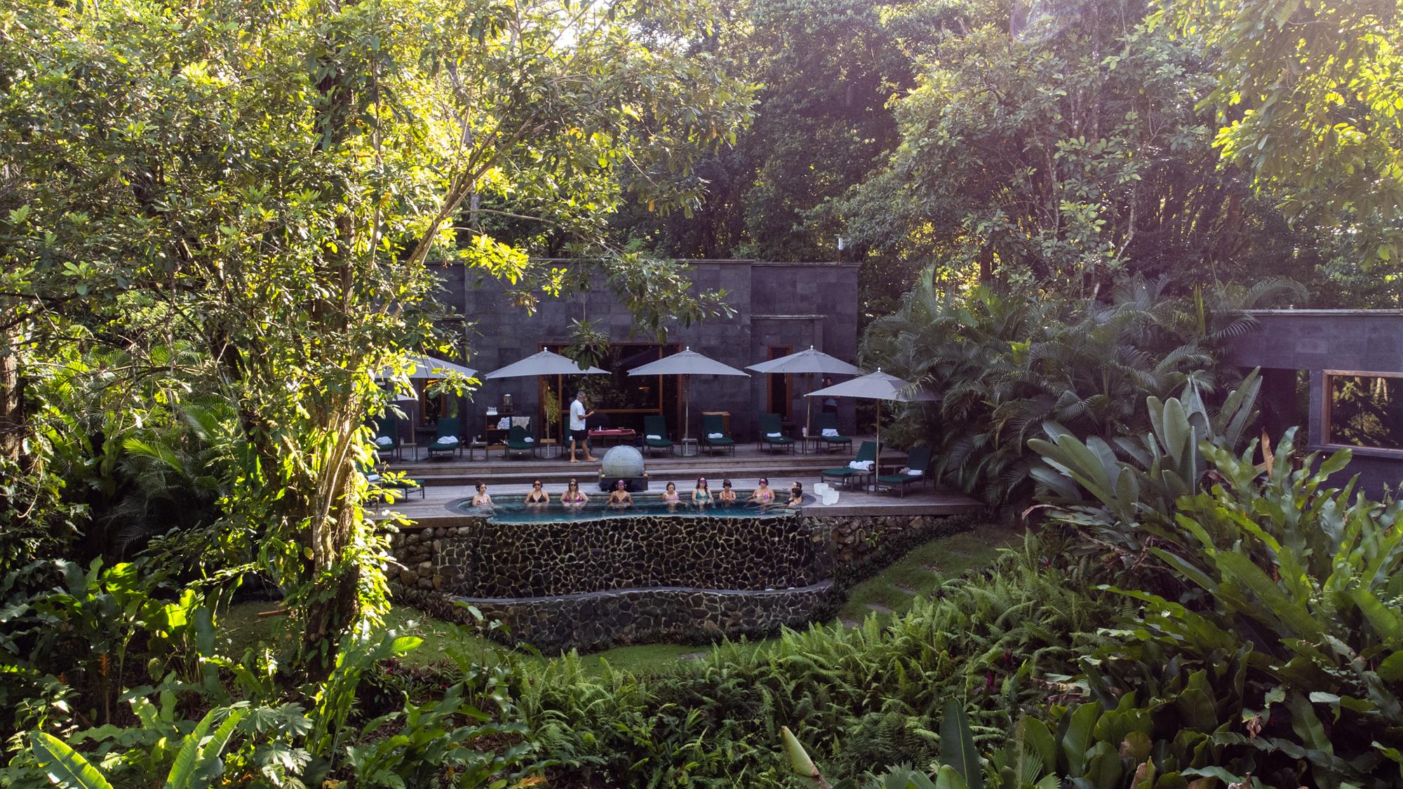 People sitting in a rectangular pool surrounded by lush tropical greenery with a modern building and umbrellas in the background.