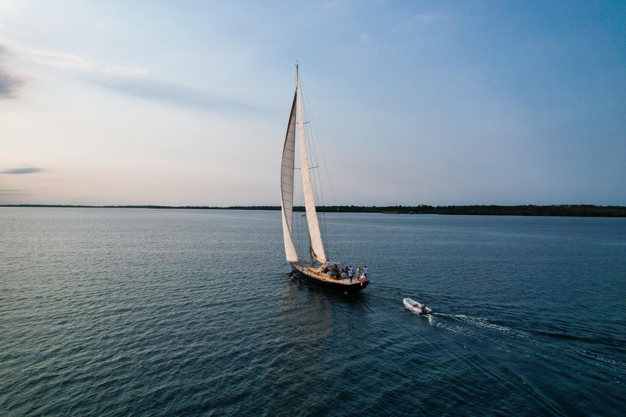 Sailboat with raised sails on calm water near a forested shoreline at dusk, followed by a small motorized dinghy.