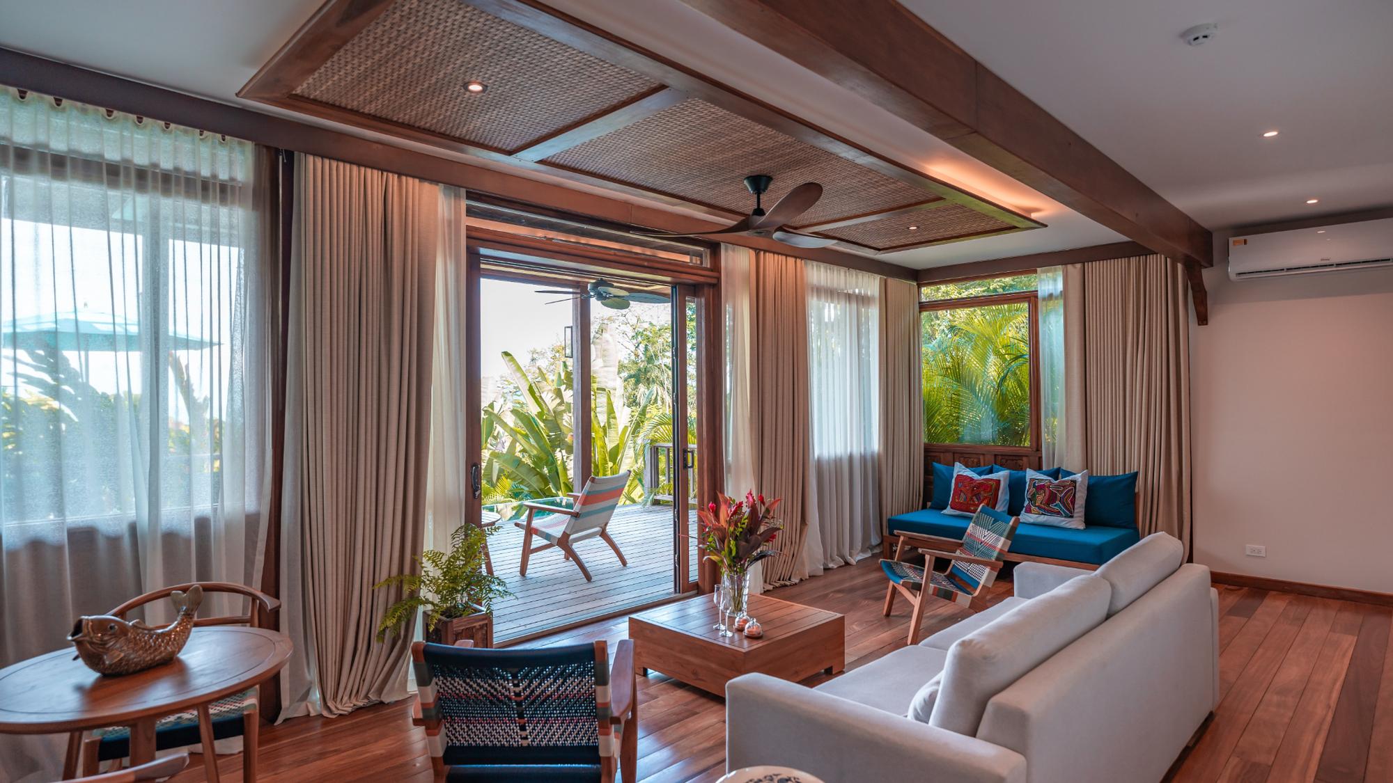 Modern living room with wood flooring, light gray sofa, blue cushioned seating, wooden coffee table, and a door opening to an outdoor deck with greenery.