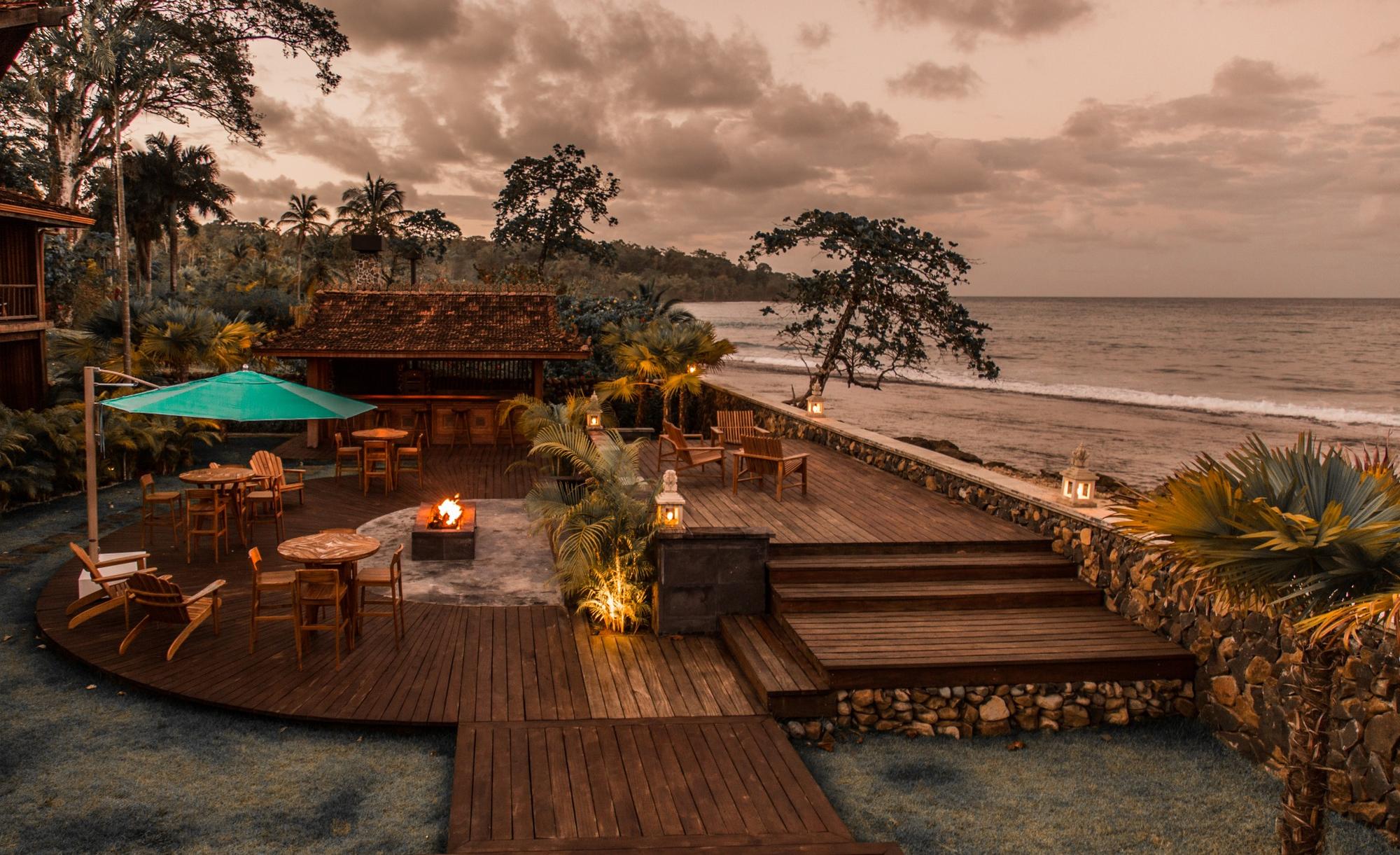 Cozy beachside wooden deck with tables, chairs, a fire pit, and a green umbrella overlooking the ocean at dusk.