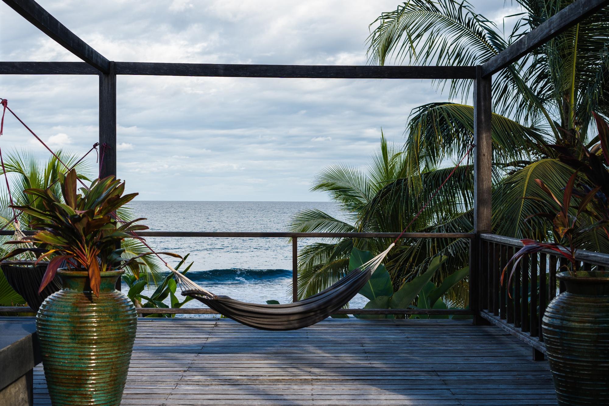 Empty hammock tied between wooden posts on a deck overlooking the ocean with palm trees and potted plants.