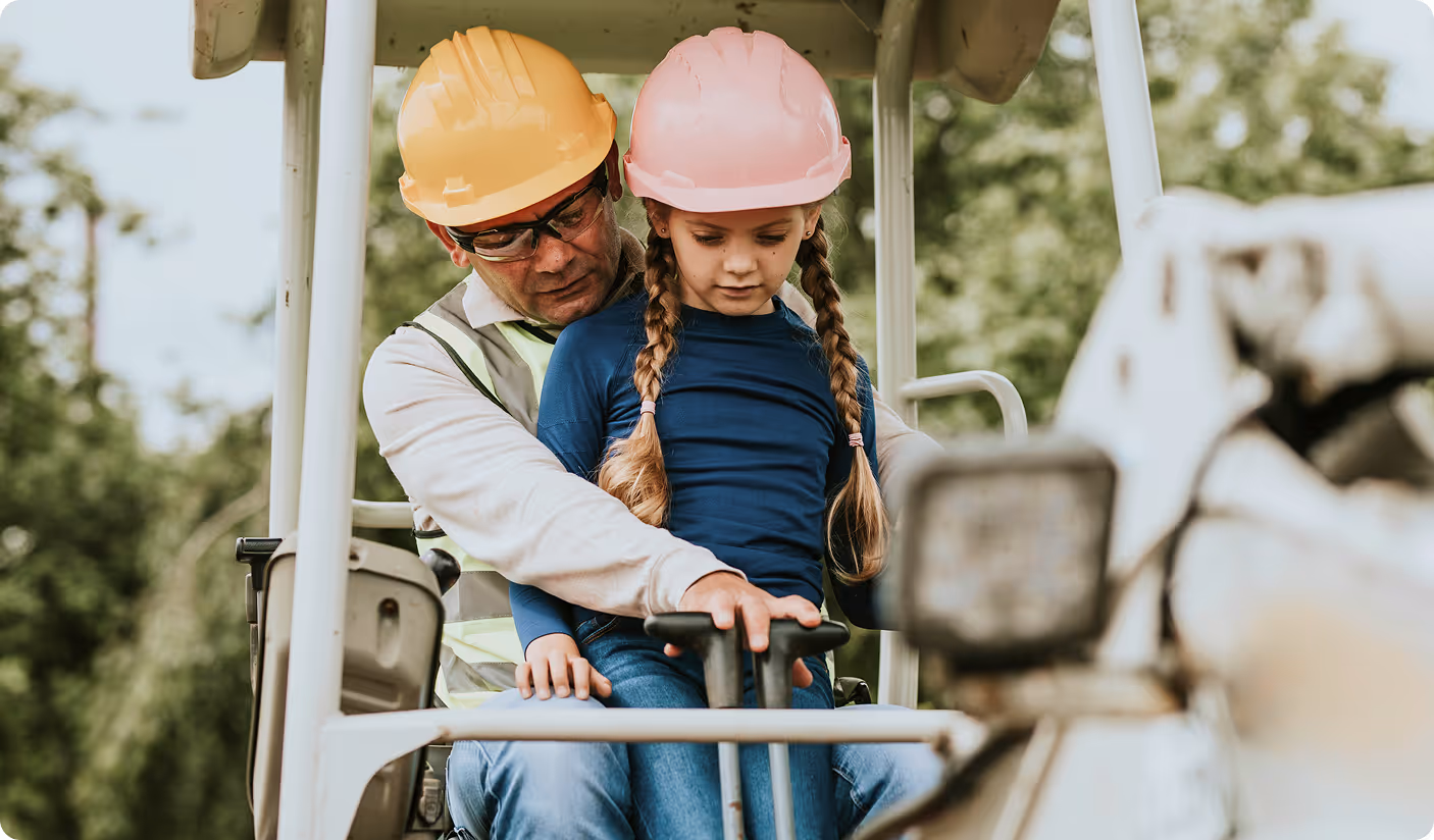 Man wearing yellow hard hat and safety glasses show a young girl in a pink hard hat construction equipment.
