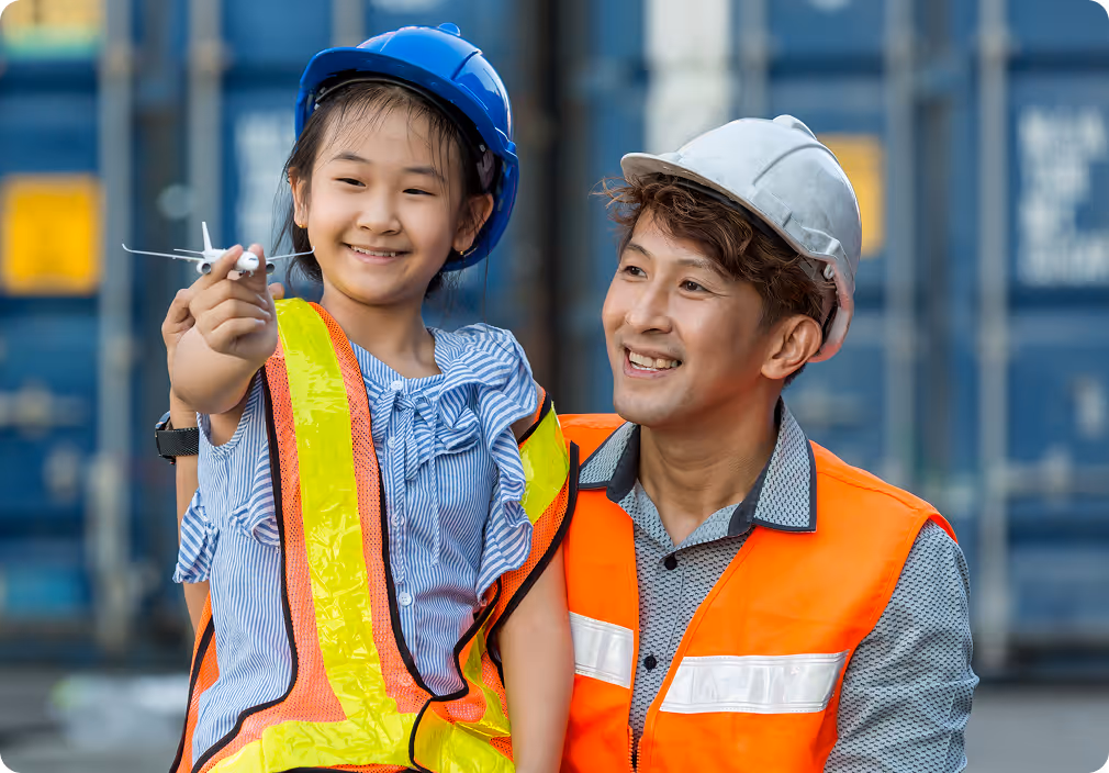 Smiling man and girl wearing safety helmets and vests, girl holding a toy airplane at a shipping container yard.