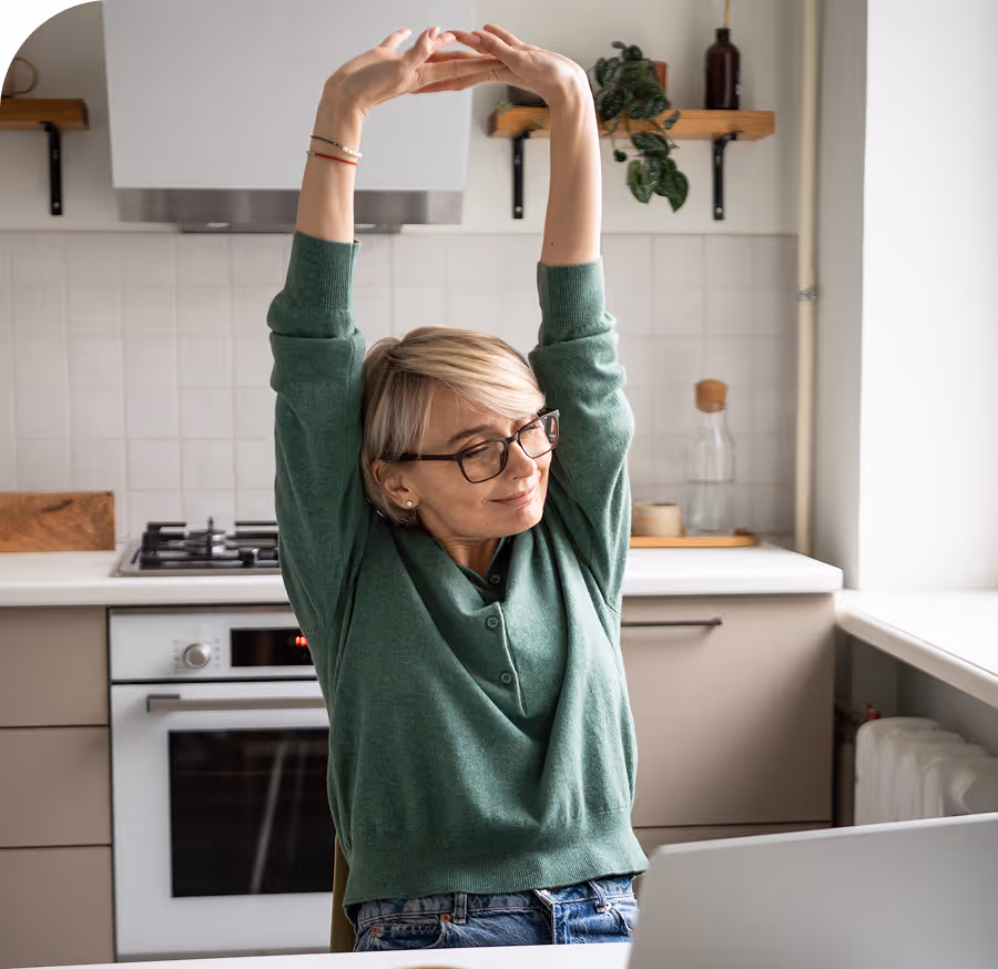 Middle-aged woman in glasses stretching her arms above her head while sitting in a kitchen with a laptop in front of her.