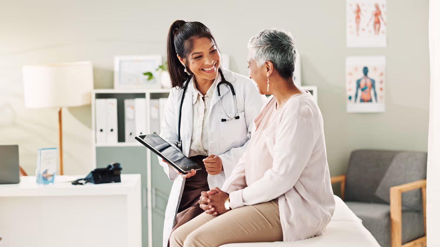 Female doctor with a stethoscope showing a tablet to a smiling older female patient in a medical office.