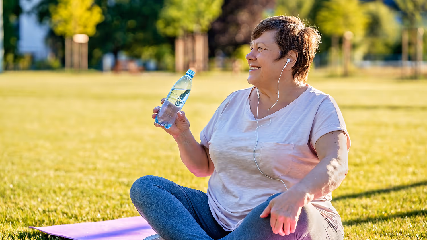 Smiling woman sitting cross-legged on a yoga mat in a park, holding a water bottle and wearing earphones.