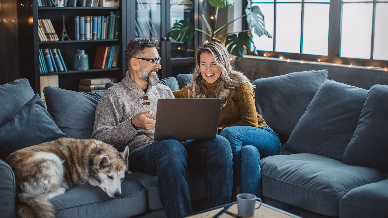Smiling couple sitting on a couch looking at a laptop with a dog resting beside them.