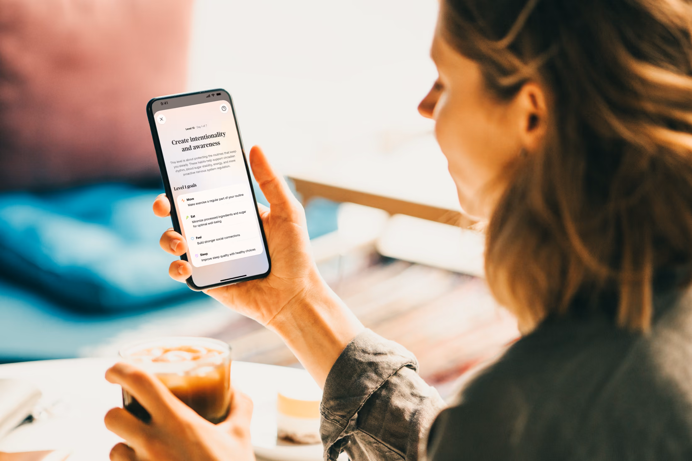 Person holding a smartphone displaying health goals and drinking an iced coffee indoors.