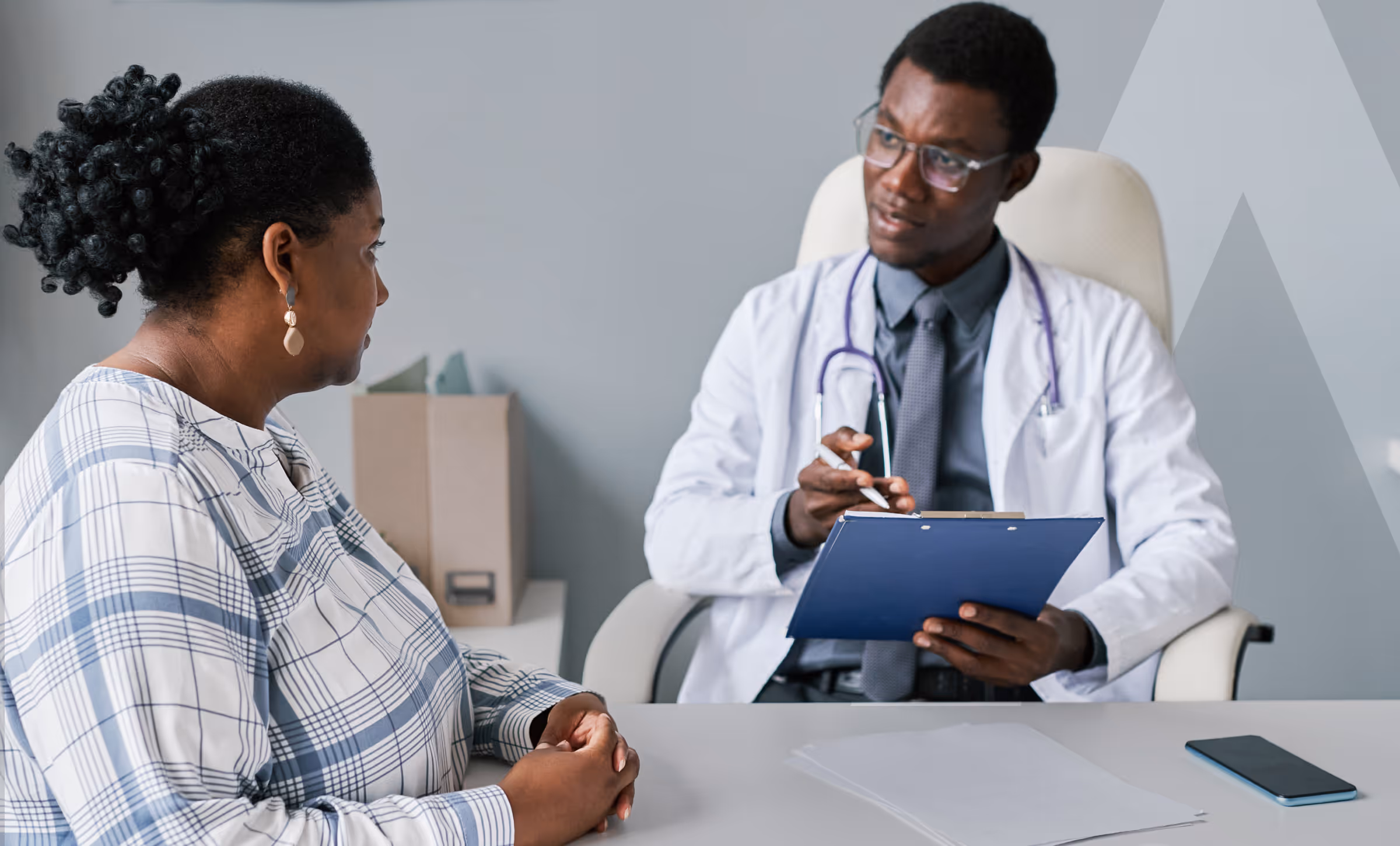 A female patient in a plaid shirt consults attentively with a male doctor with a stethoscope and clipboard in a medical office.