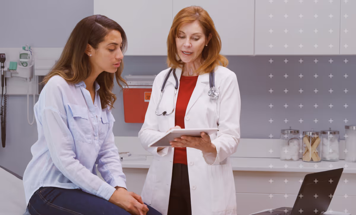 Female doctor in a white coat with a stethoscope consulting a seated female patient in a medical exam room.