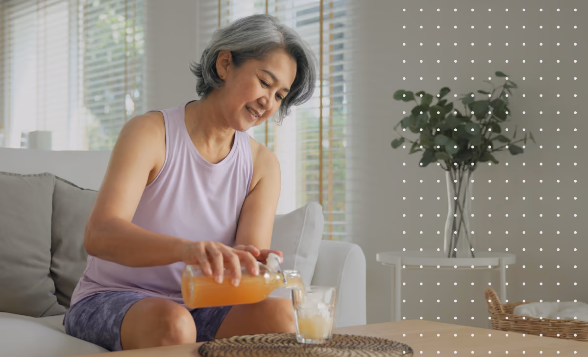 Smiling older woman in purple shirt pouring juice into a glass while sitting on a sofa.
