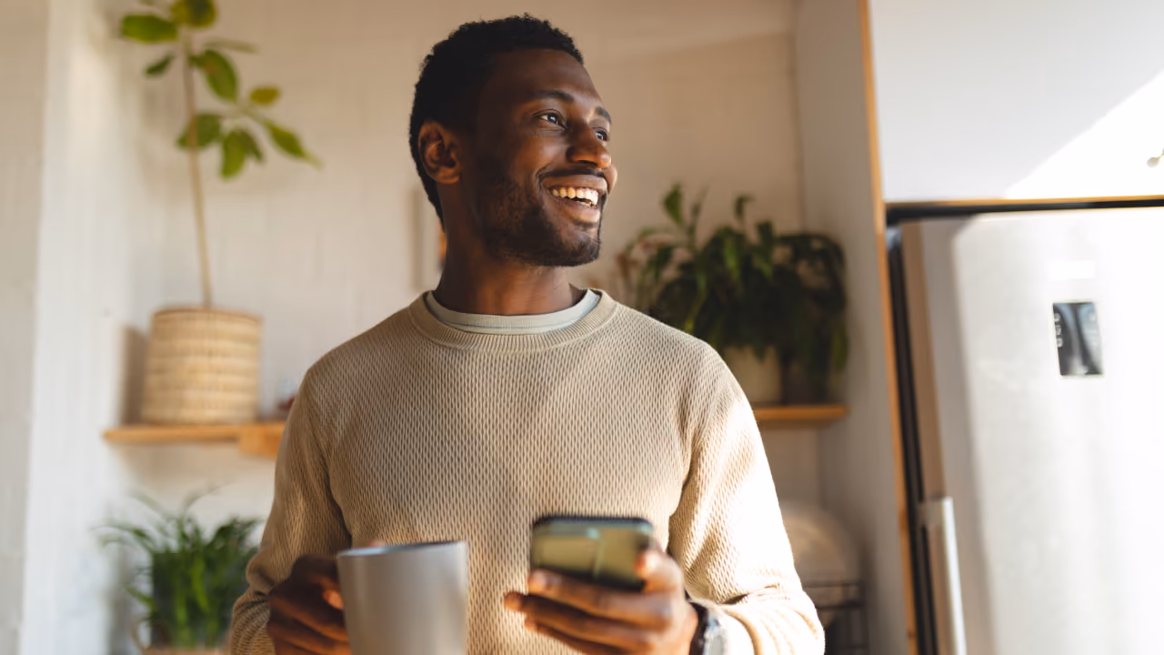 Smiling man holding a phone and a cup in a bright kitchen with plants and a refrigerator.