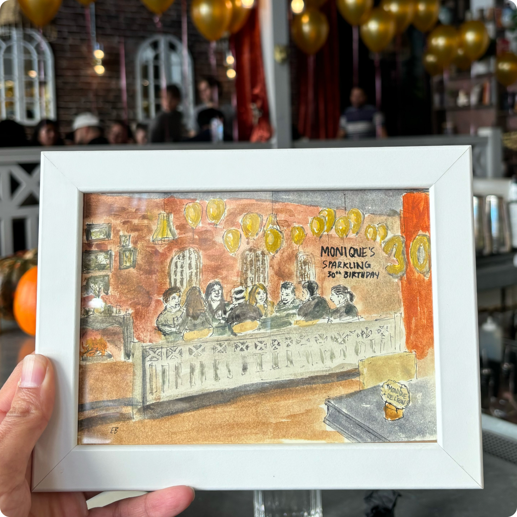 Hand holding a white-framed watercolor painting of people celebrating with balloons and a banner reading 'Monique's Sparkling 30th Birthday.'