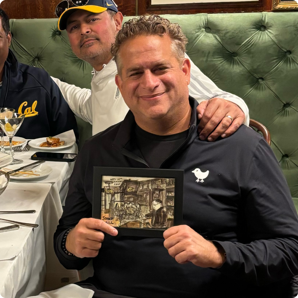 Smiling man holding a framed artwork seated at a restaurant table with another man in a white shirt and blue and yellow cap behind him.