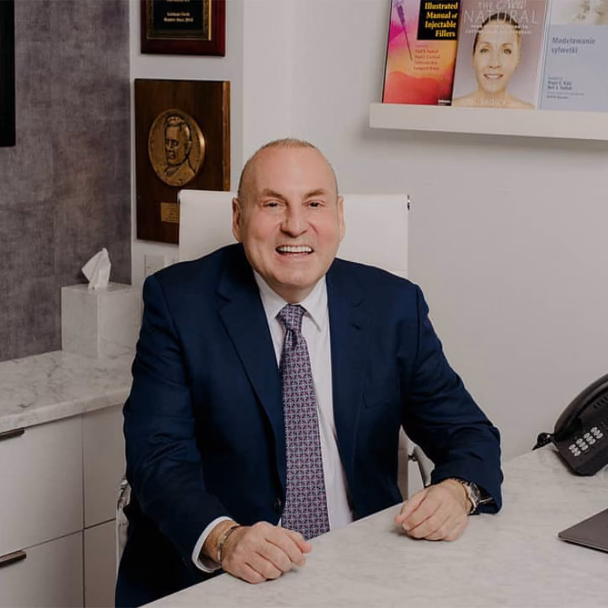 Dermatologist Dr. Neil Sadick sits in his office wearing a black jacket and smiling.