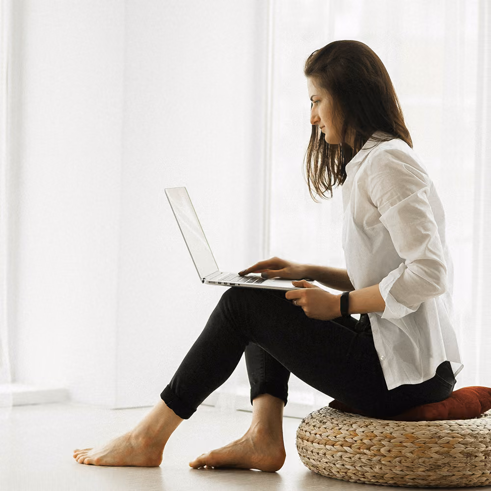 A woman sits on a wicker chair working on her laptop.