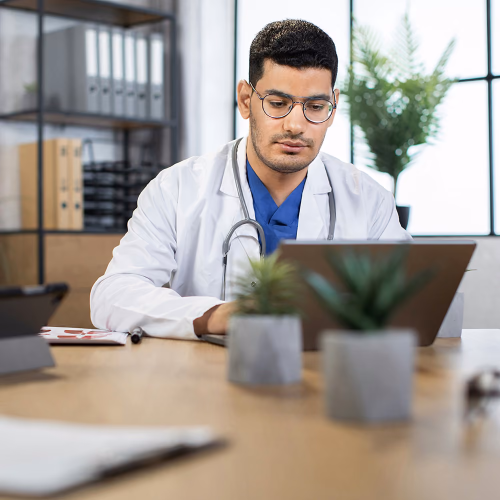 The doctor is in his office typing something on his laptop.
