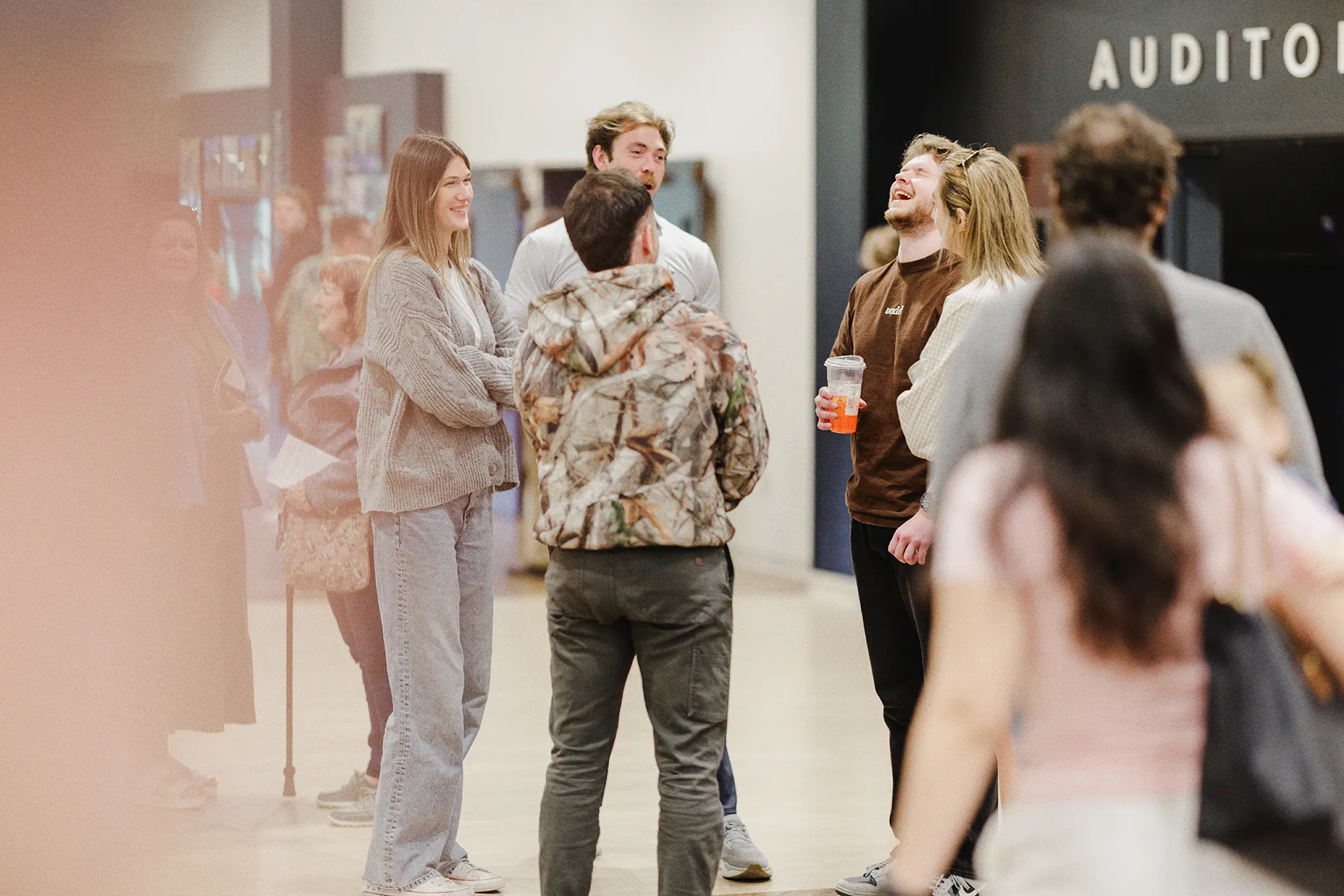 Young adults laughing and talking in the church lobby before the service, with the auditorium entrance visible in the background.