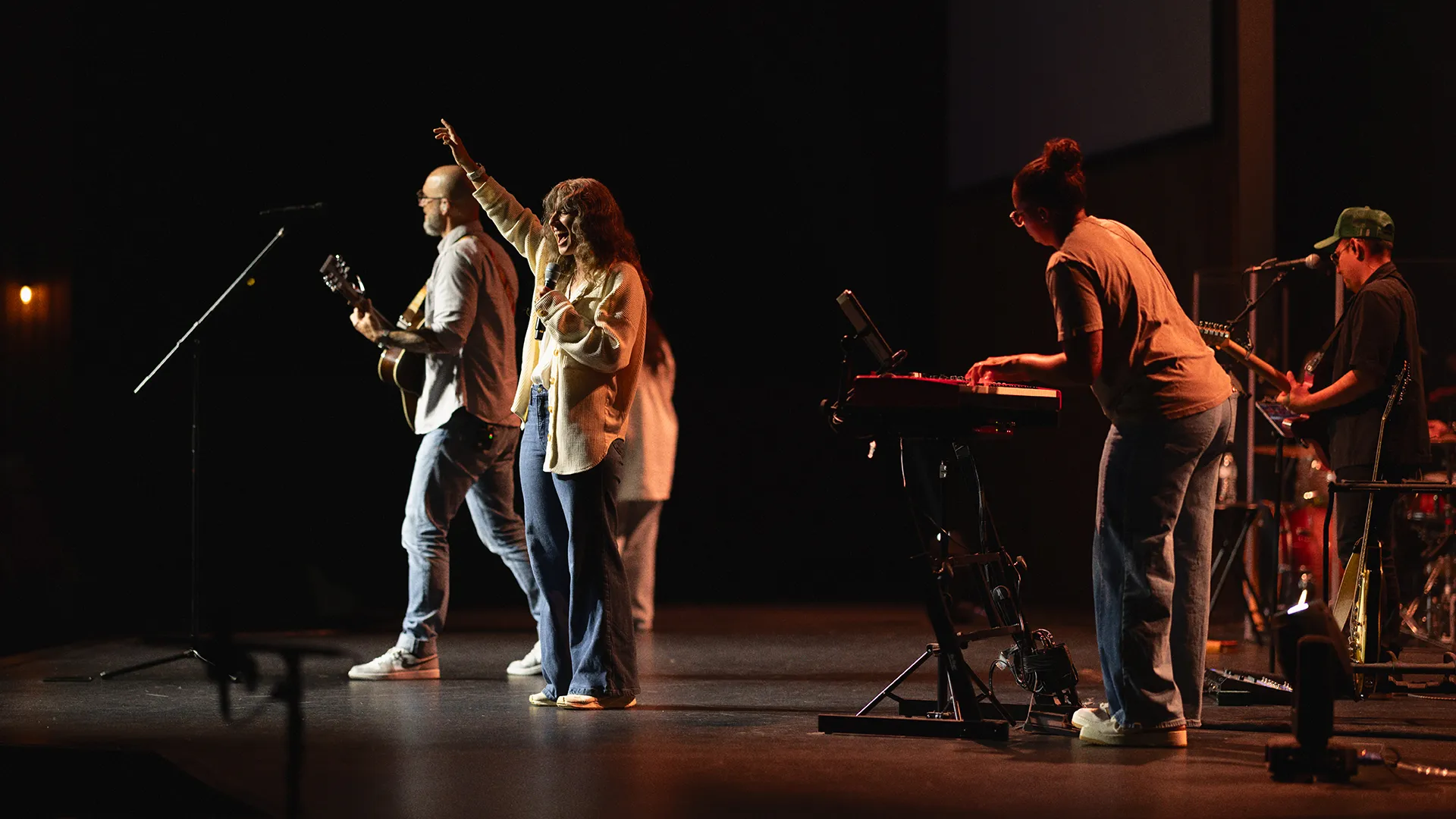 Worship band performing on stage during a Sunday service, dimly lit with a vocalist raising her hand.