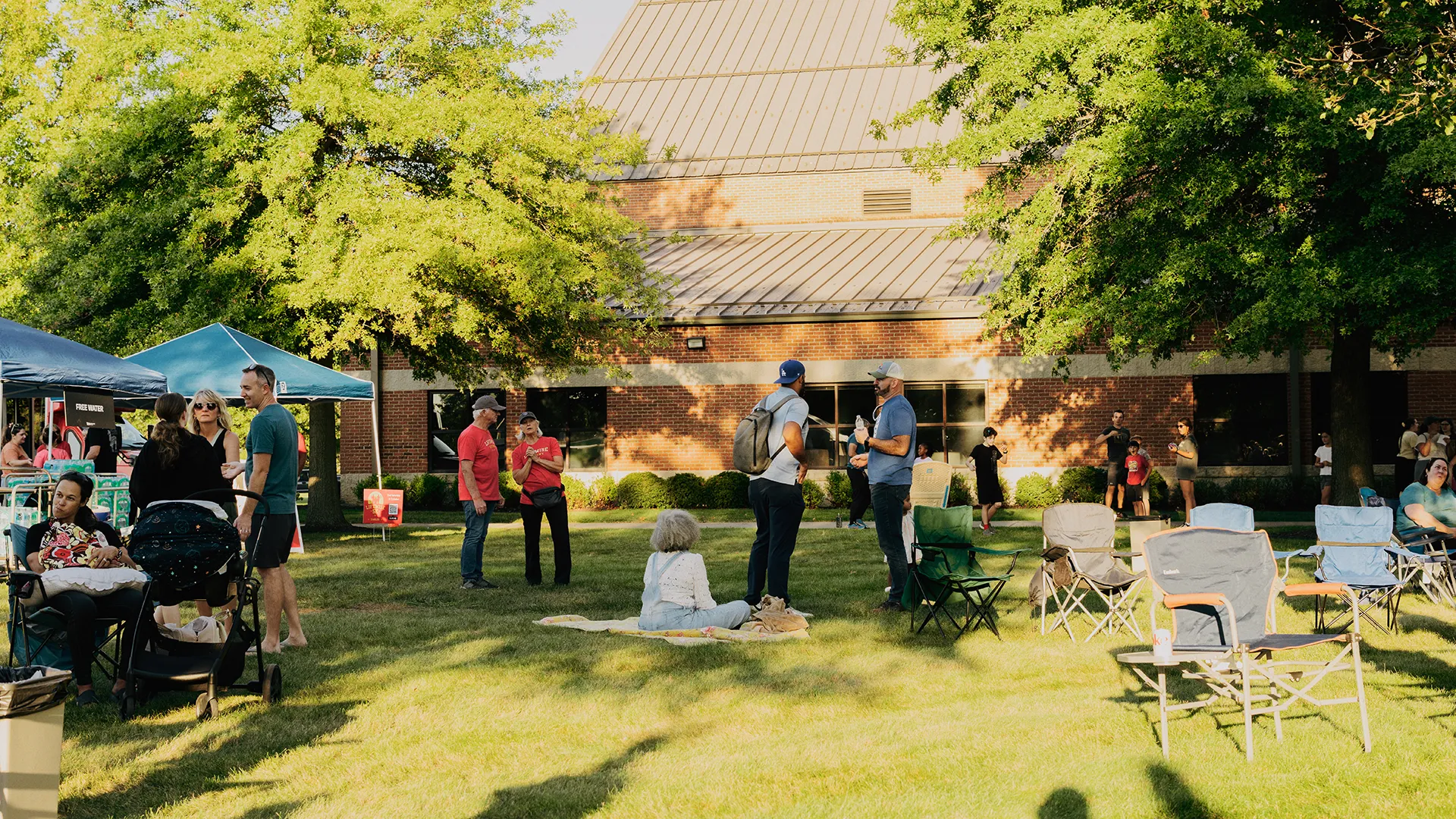 People gathered on the church lawn for a summer evening outdoor event, sitting in camp chairs and on blankets under shade trees.