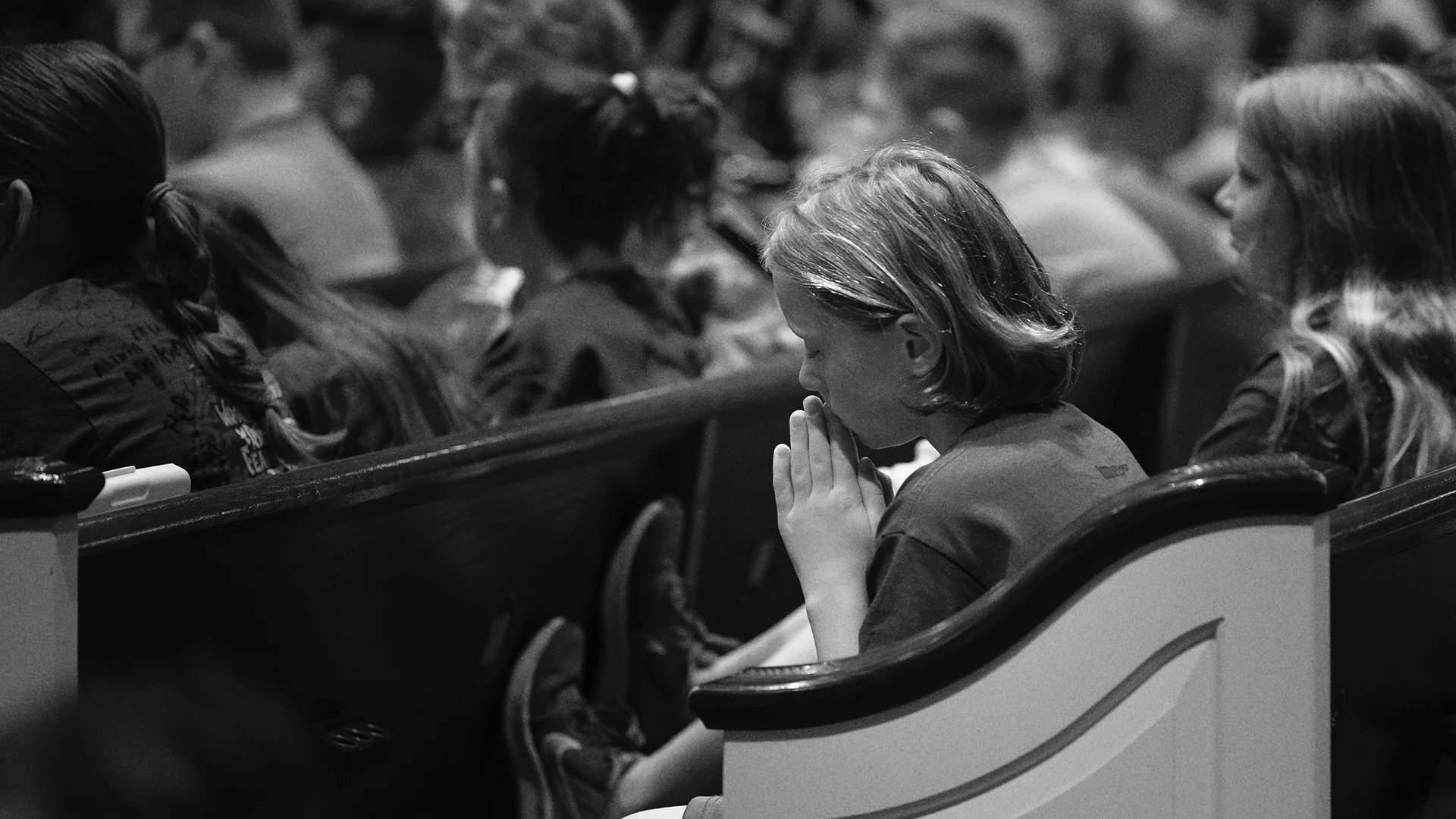 A child prays with hands folded during VBS at Northwest Bible Church, surrounded by a full room of kids