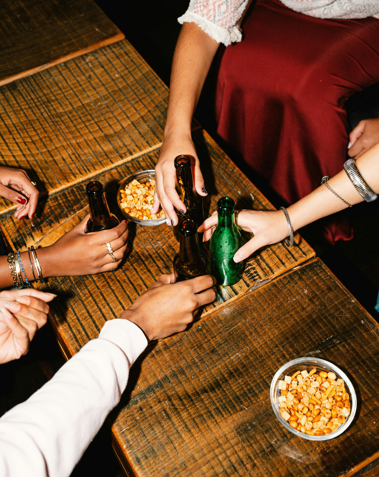 Four people clinking beer bottles over a wooden table with snacks in glass bowls.