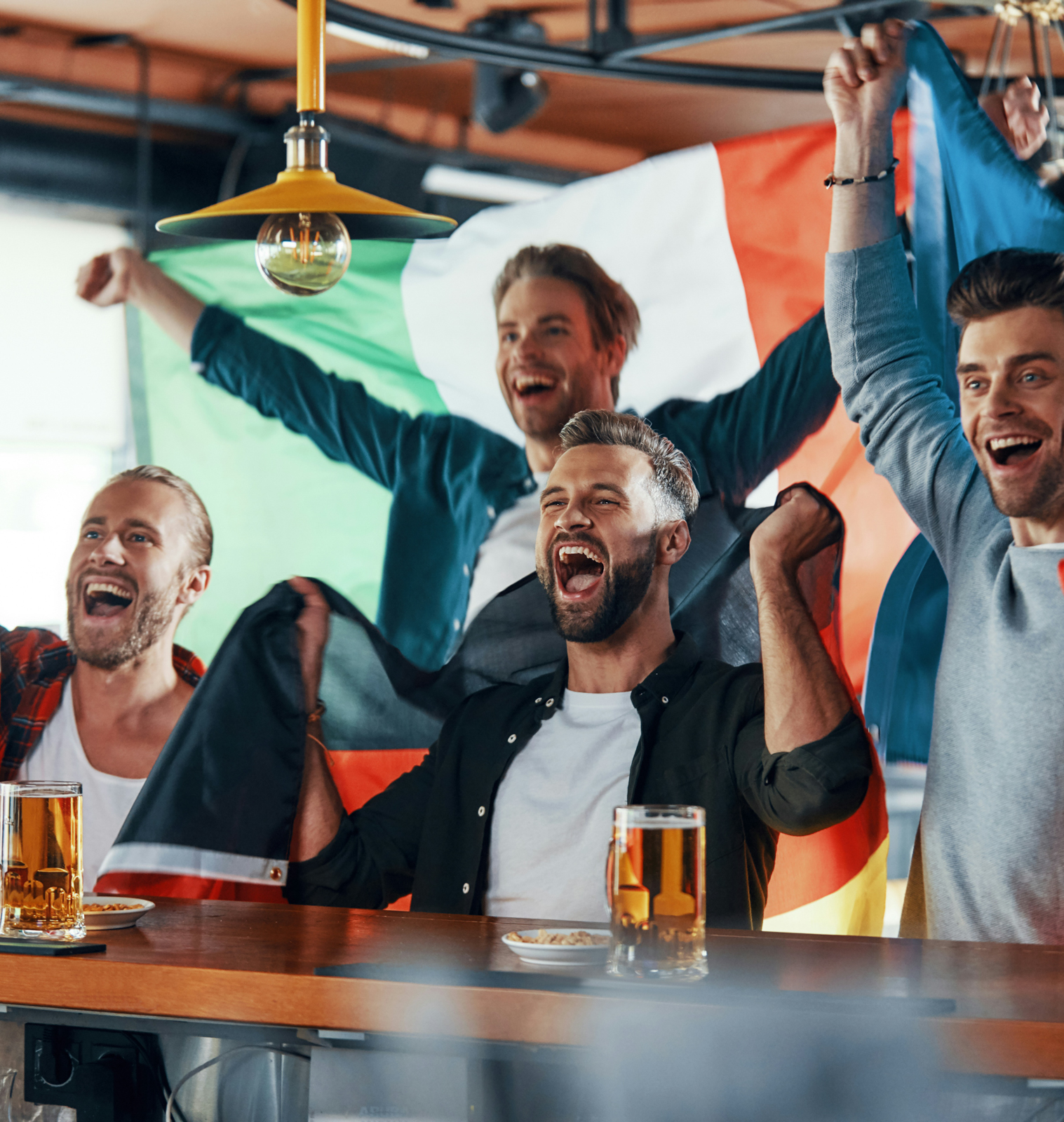 Four men cheering at a bar holding UK, Germany, Ireland, and France flags with beer mugs on the counter.