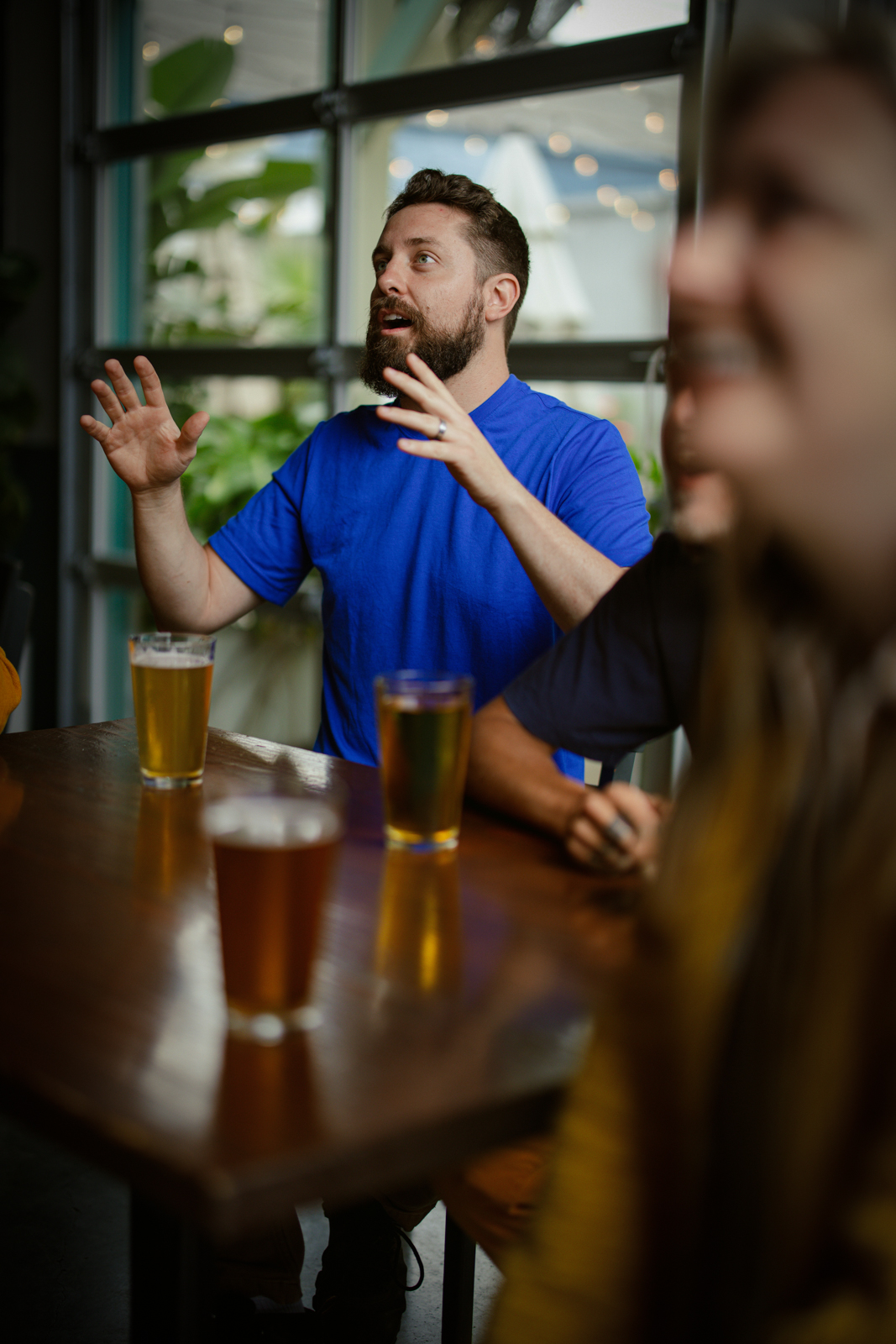 A man with a beard and blue shirt is sitting at a table with two beers.