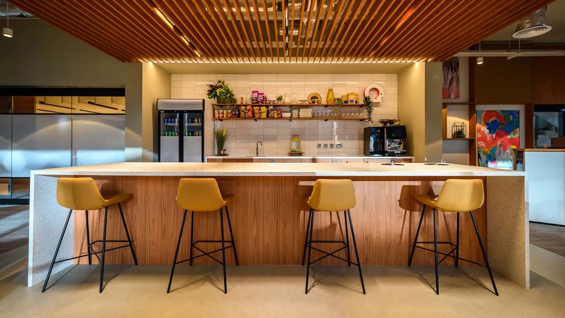 Modern coworking kitchen with a large island, bar stools, wooden ceiling slats, and open shelving.