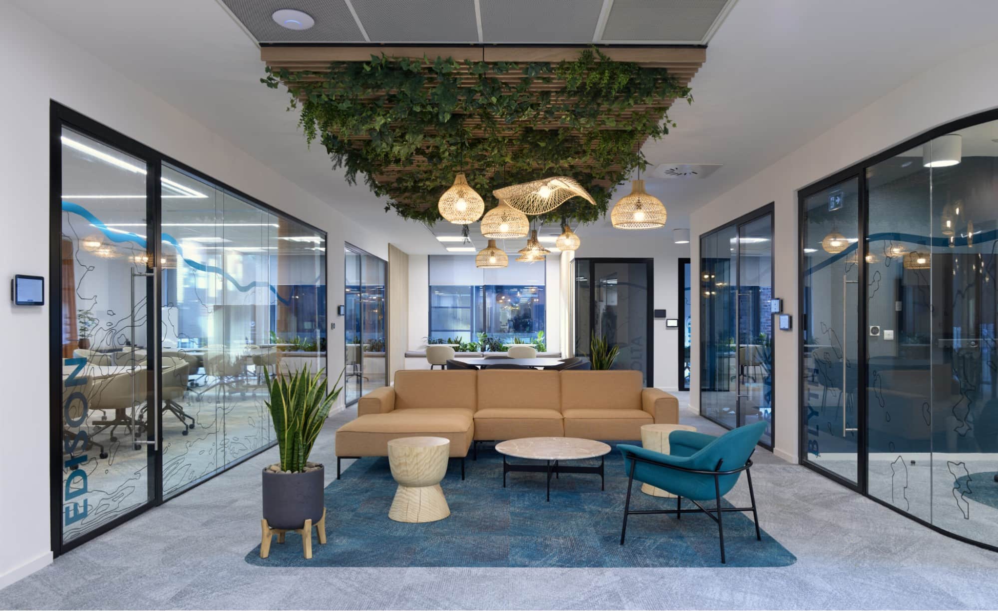 Contemporary coworking lounge with a tan sofa, modern seating, hanging woven lights, and glass meeting rooms surrounded by greenery.