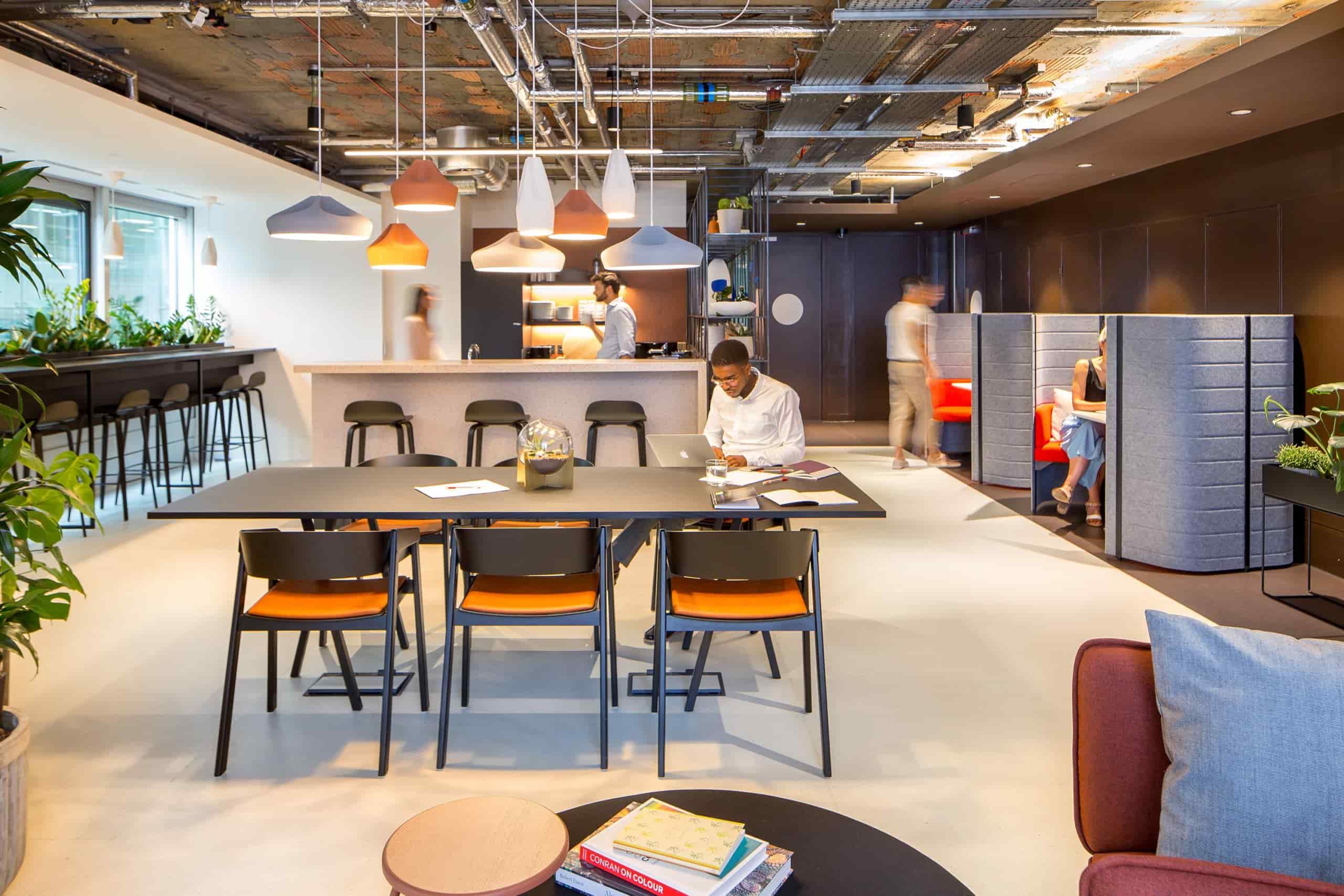 Vibrant coworking lounge with shared tables, private work booths, a communal bar area, and exposed industrial ceilings.