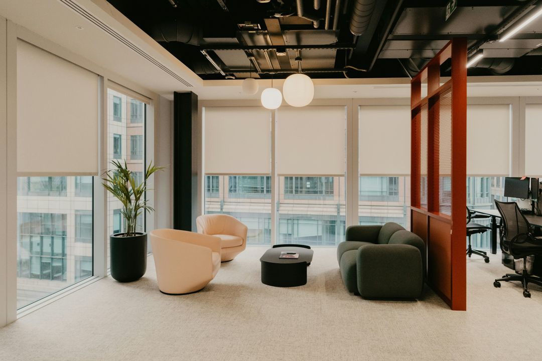 Modern office lounge area with contemporary armchairs, a low black coffee table, large windows, and exposed ceiling features.