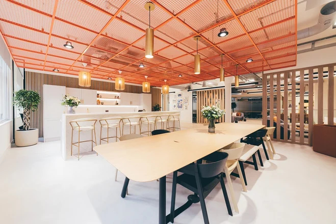 Bright modern office breakout area with a long communal table, bar seating, warm pendant lighting, and an orange mesh ceiling feature.