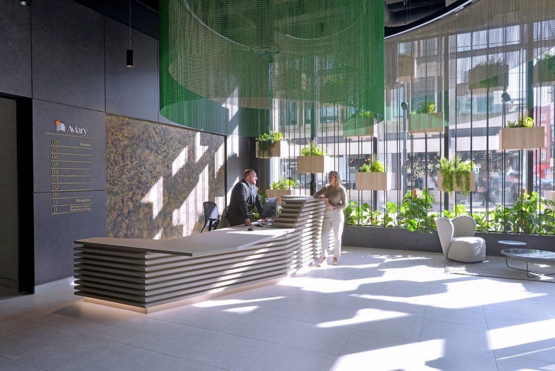 Modern office reception area with sculpted front desk, floor-to-ceiling windows, hanging green mesh feature, and lush indoor planting at Aviary Manchester.