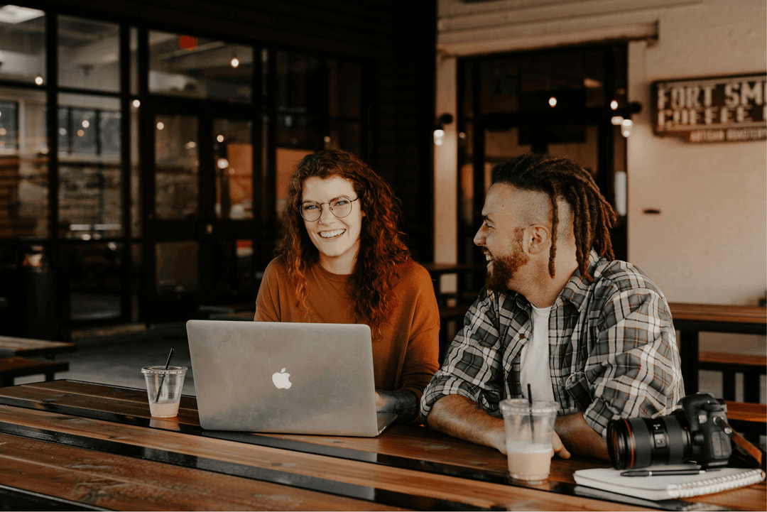 Two people collaborating on a laptop in a relaxed workspace with drinks and a camera on the table.