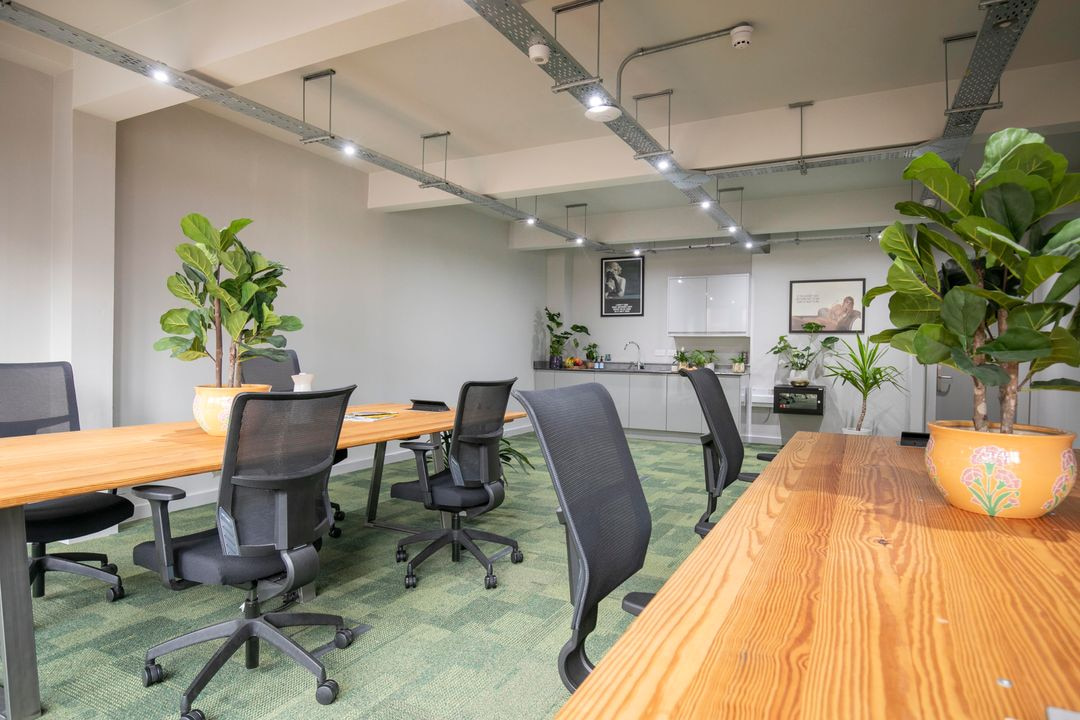 Modern serviced office with large wooden coworking desks, ergonomic mesh chairs, greenery, and a stylish kitchenette.