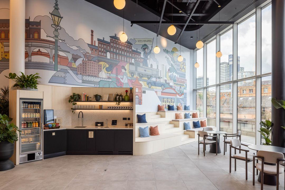 Modern office breakout space with tiered seating, large city-view windows, vibrant wall mural and a self-serve kitchen area.